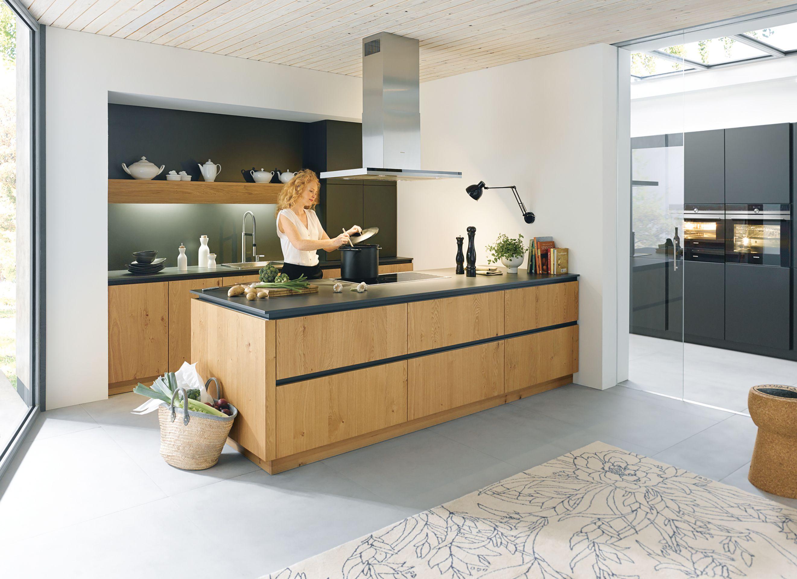 Moderne, helle Einbauküche mit Kochinsel aus Holz und dunkler Arbeitsplatte, eine Frau kocht entspannt, im Hintergrund Regale und Geräte – Beispiel für hochwertige Küchen in Füssen und dem Allgäu.