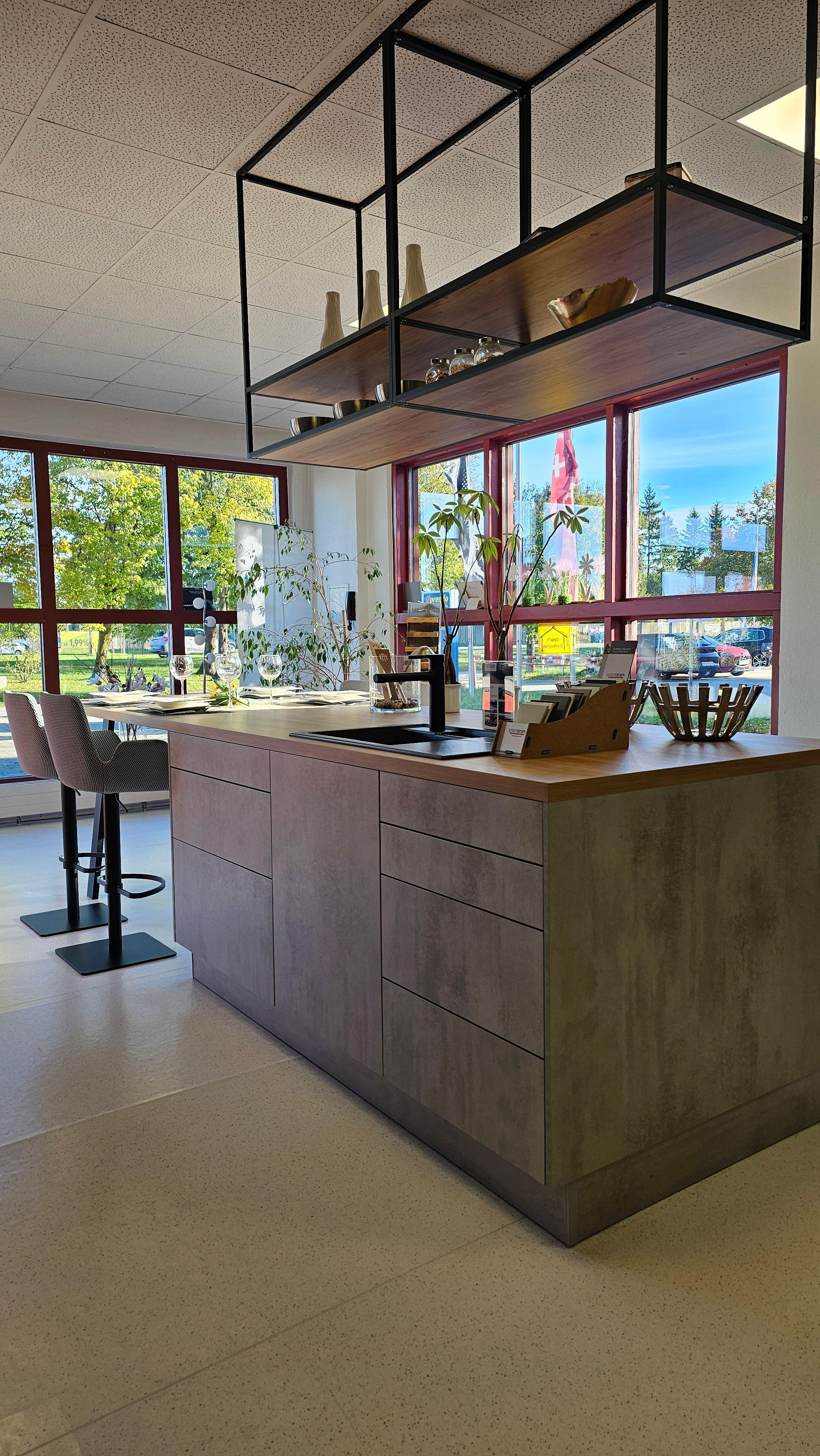 Moderne Kochinsel mit Betonoptik-Fronten, schwarzer Armatur und offenem Deckenregal vor großen Fenstern – zeitgemäße Küchen in Füssen und dem Allgäu.