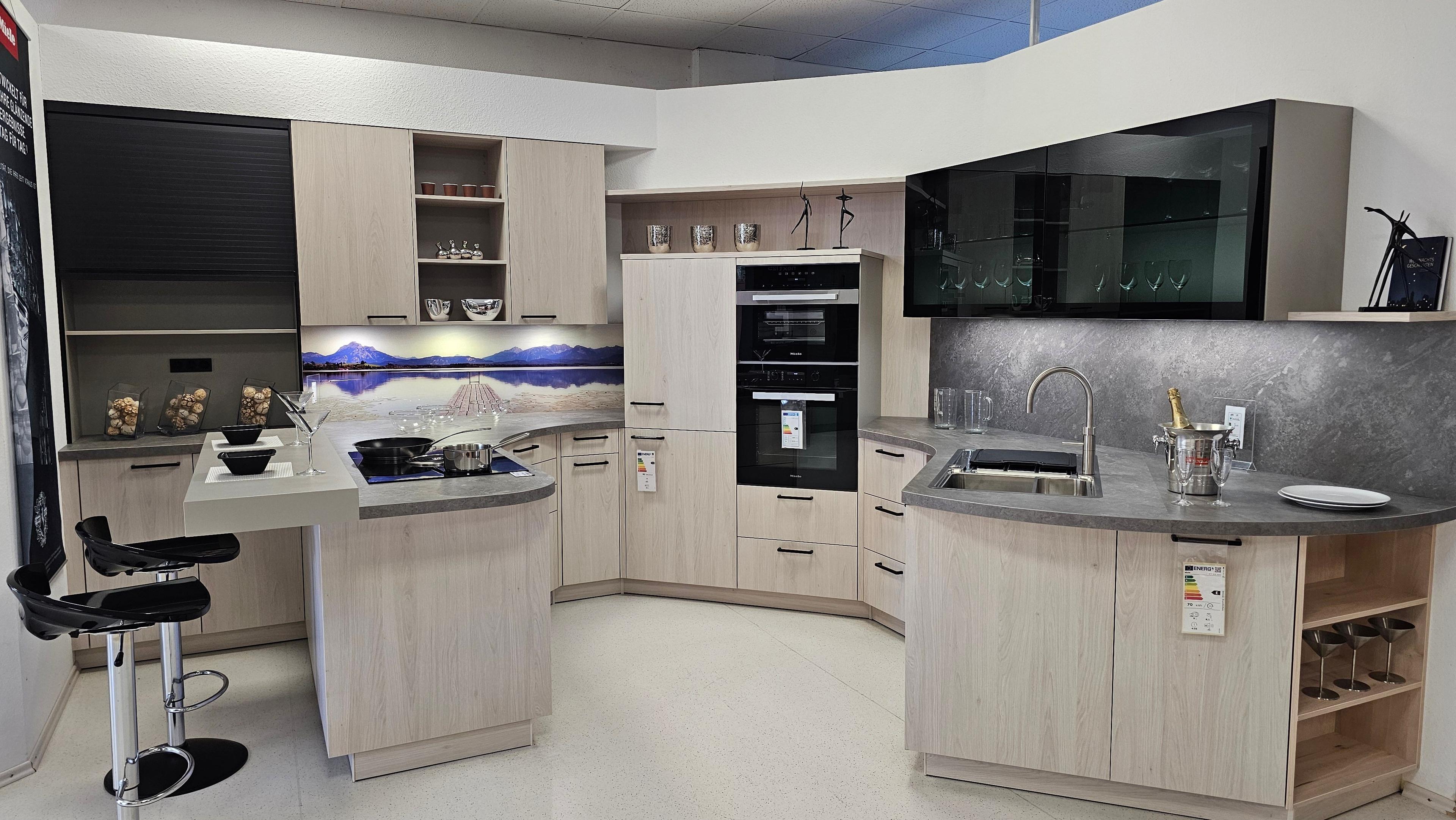 U-förmige Küche mit hellen Holzfronten, Glasoberschränken, Tresen und Geräten auf Augenhöhe – vielseitige Küchen in Füssen und dem Allgäu.