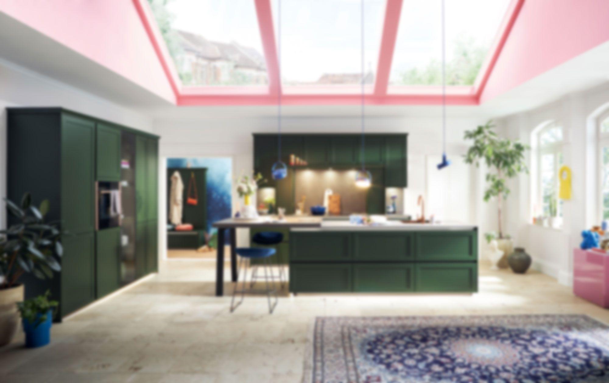 Moderne Küche in dunklem Grün mit großer Kochinsel, offenen Regalen und farbenfrohem Interior unter Glasdach – stilvolle Schüller Küchen im Allgäu für individuelles Design und hochwertige Ausstattung.