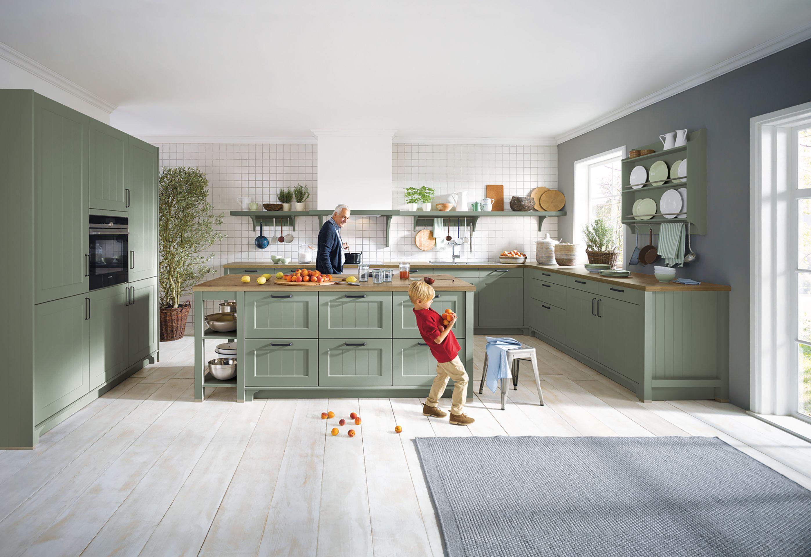 Großzügige grüne Landhausküche mit Kochinsel, offenen Regalen und Holzarbeitsplatten, Kind spielt im Raum – familienfreundliche Küchen in Füssen und dem Allgäu.