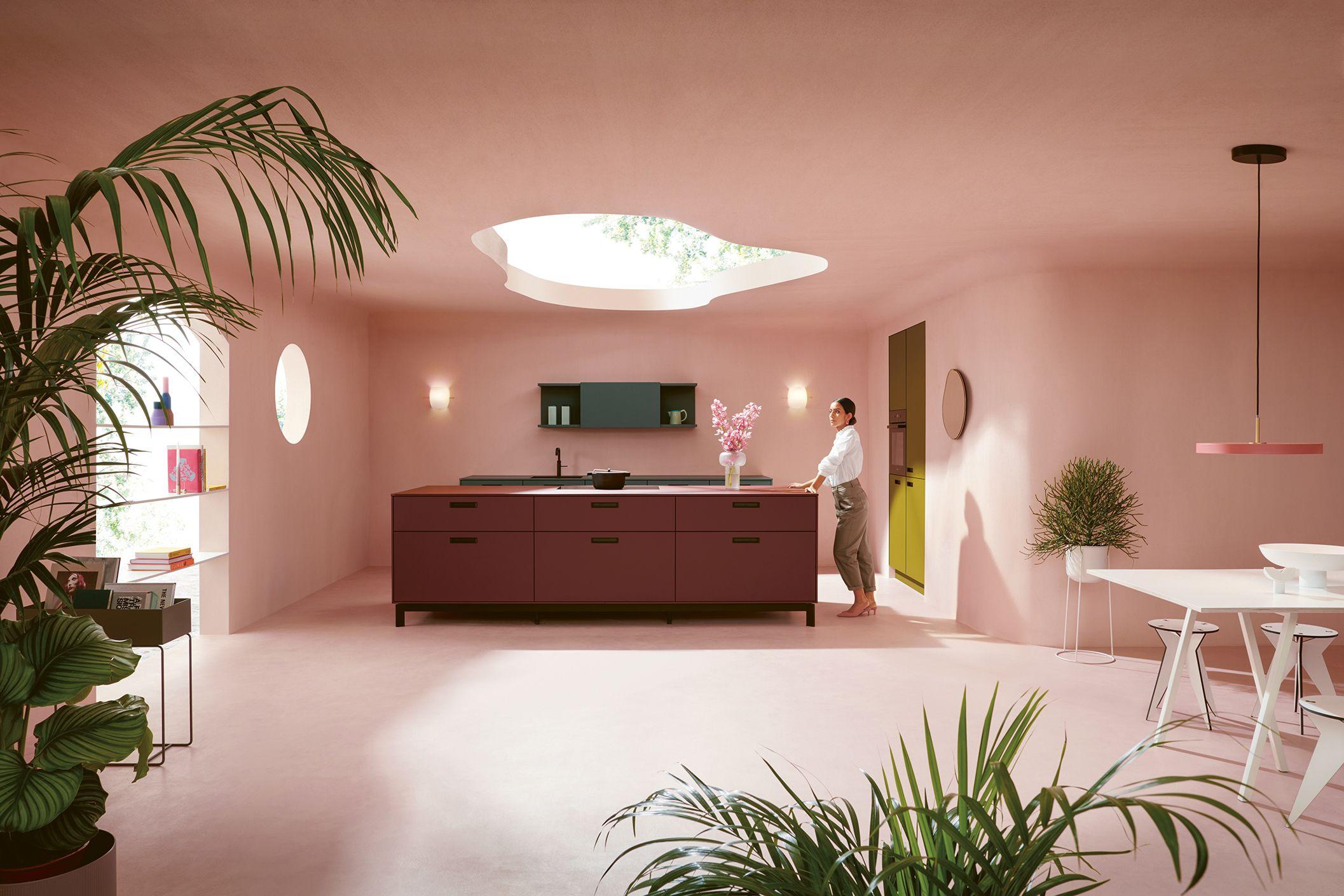 Designorientierte Küche mit großer Kochinsel in Bordeauxrot, offenen Regalen und Oberlicht in einem modernen rosa Raum, eine Person lehnt entspannt daran – stilvolle Küchen in Füssen und dem Allgäu.