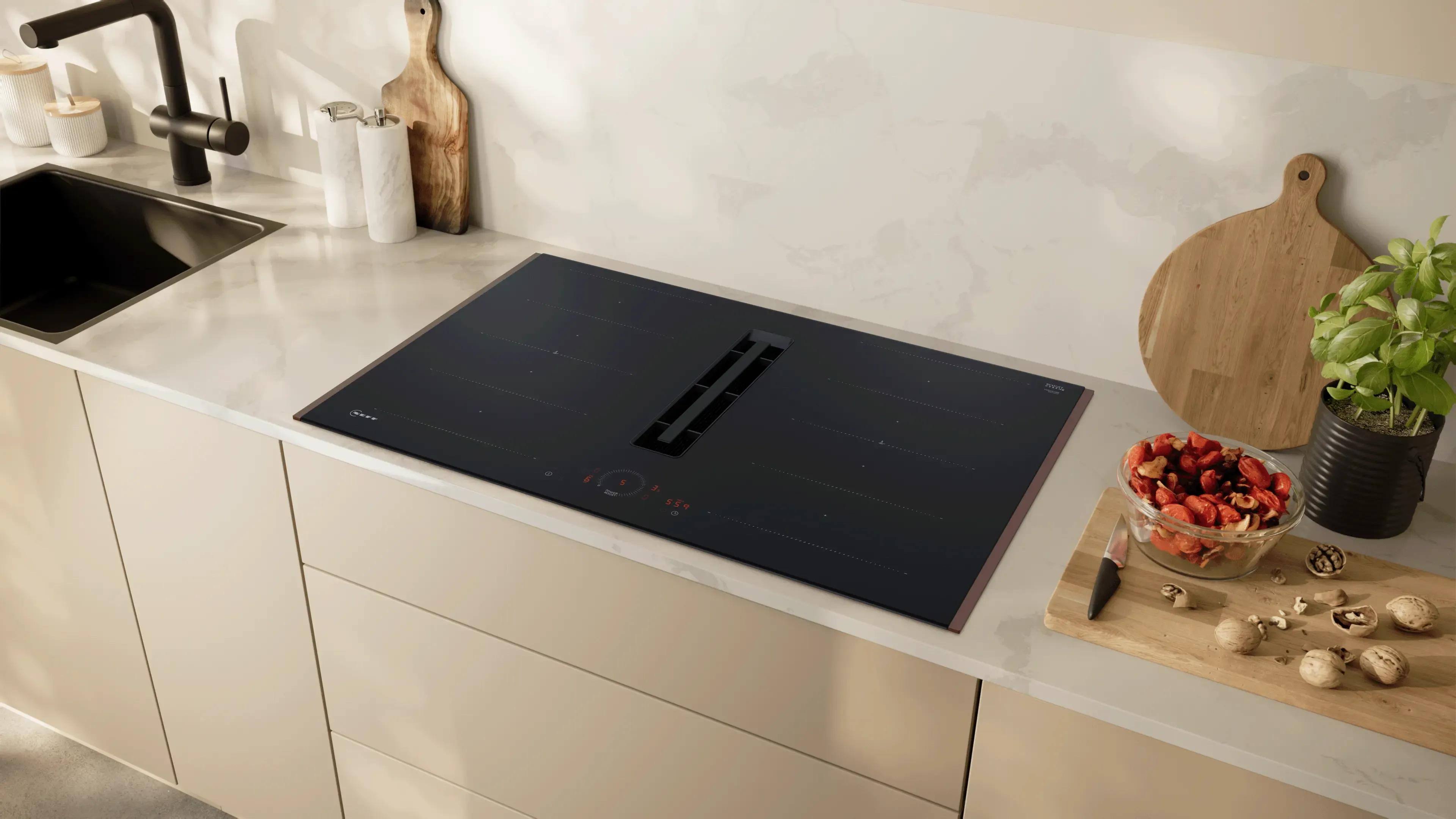 Küchenarbeitsplatte mit integriertem schwarzen Neff Flex Design Kochfeld, links Spüle mit Armatur, rechts Schneidebrett, Schüssel mit Gemüse und Kräutertopf.