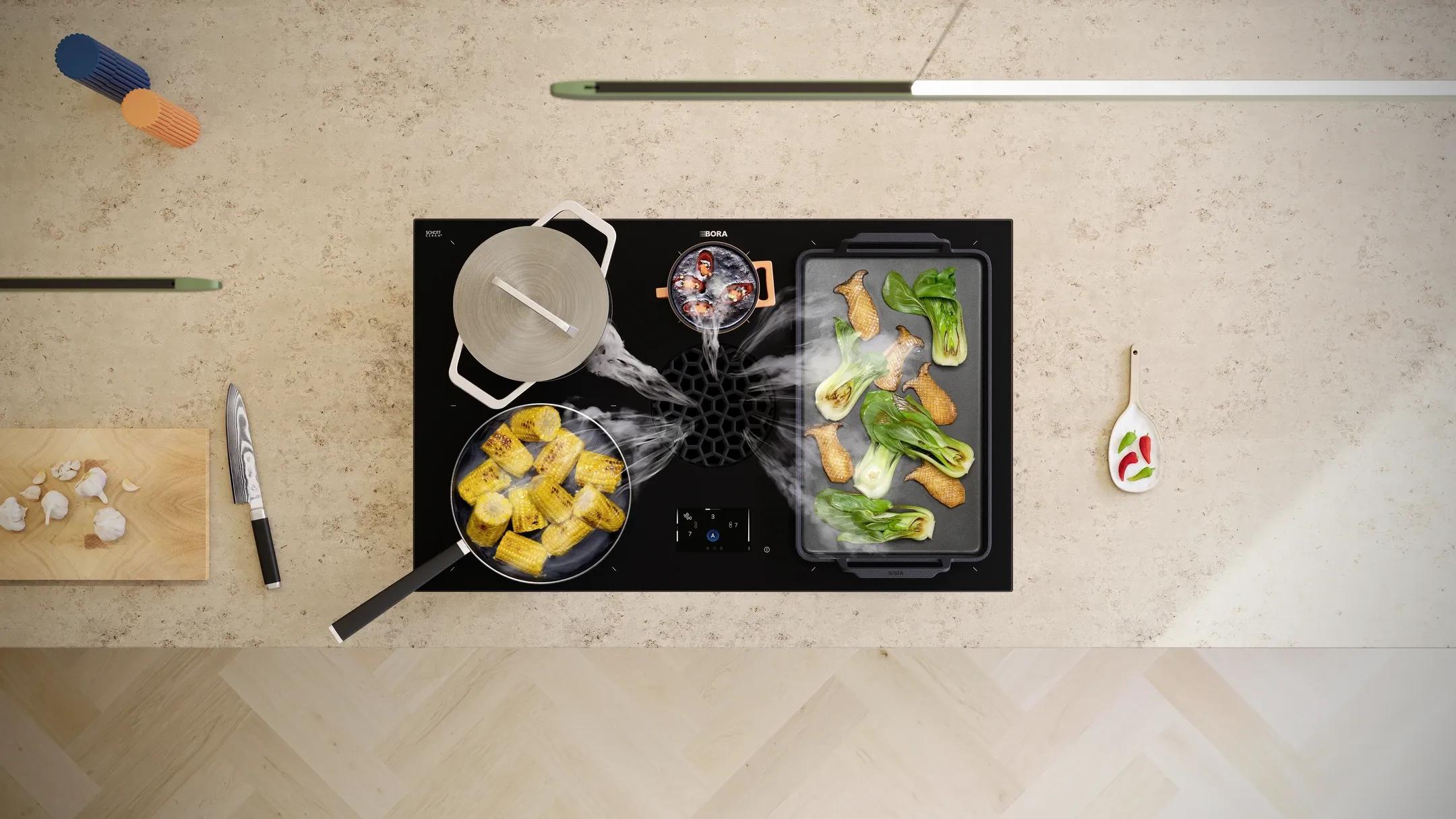 Draufsicht auf ein Bora FK06 X-Pure Max Kochfeld mit Dunstabzug, auf dem Maiskolben in einem Topf und verschiedene Gemüse auf einer Grillplatte zubereitet werden, umgeben von Küchenutensilien auf einer hellen Arbeitsfläche.